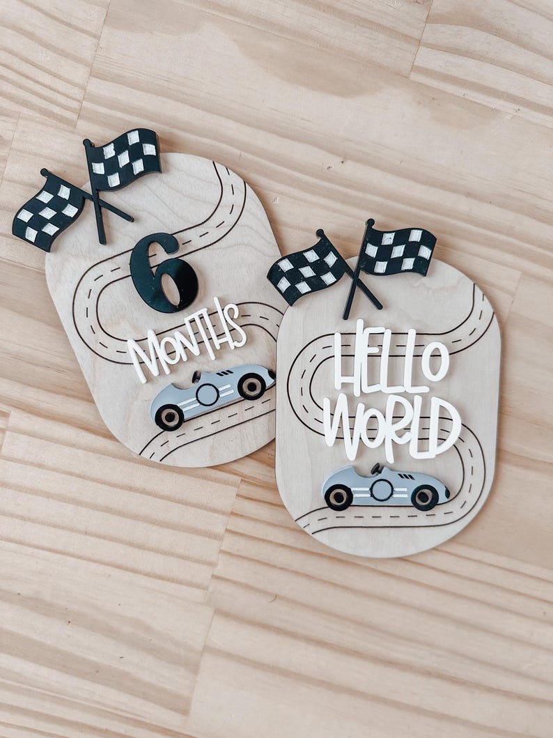 May include: Two wooden baby milestone signs featuring a race car theme. The first sign reads "6 Months" with a black number six and a black and white chequered flag. The second sign reads "Hello World" with a black and white chequered flag. Both signs have a black and white race track design and a silver race car.
