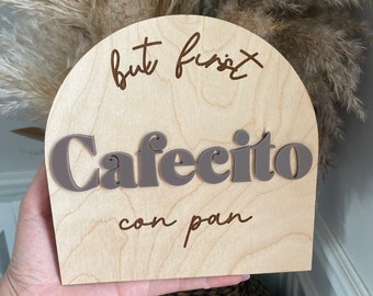 May include: Wooden sign with the text "but first, cafécito con pan" in brown lettering. The sign is shaped like an arch and is made of light wood.