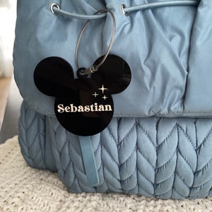 Könnte beinhalten: Ein blauer gesteppter Rucksack mit einem schwarzen Mickey-Mouse-förmigen Anhänger, auf dem "Sebastian" in goldenen Buchstaben steht.