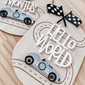 May include: Two wooden baby milestone signs with a race car theme. The first sign says "6 Months" and features a black number 6 and a grey race car. The second sign says "Hello World" and features a chequered flag and a grey race car.