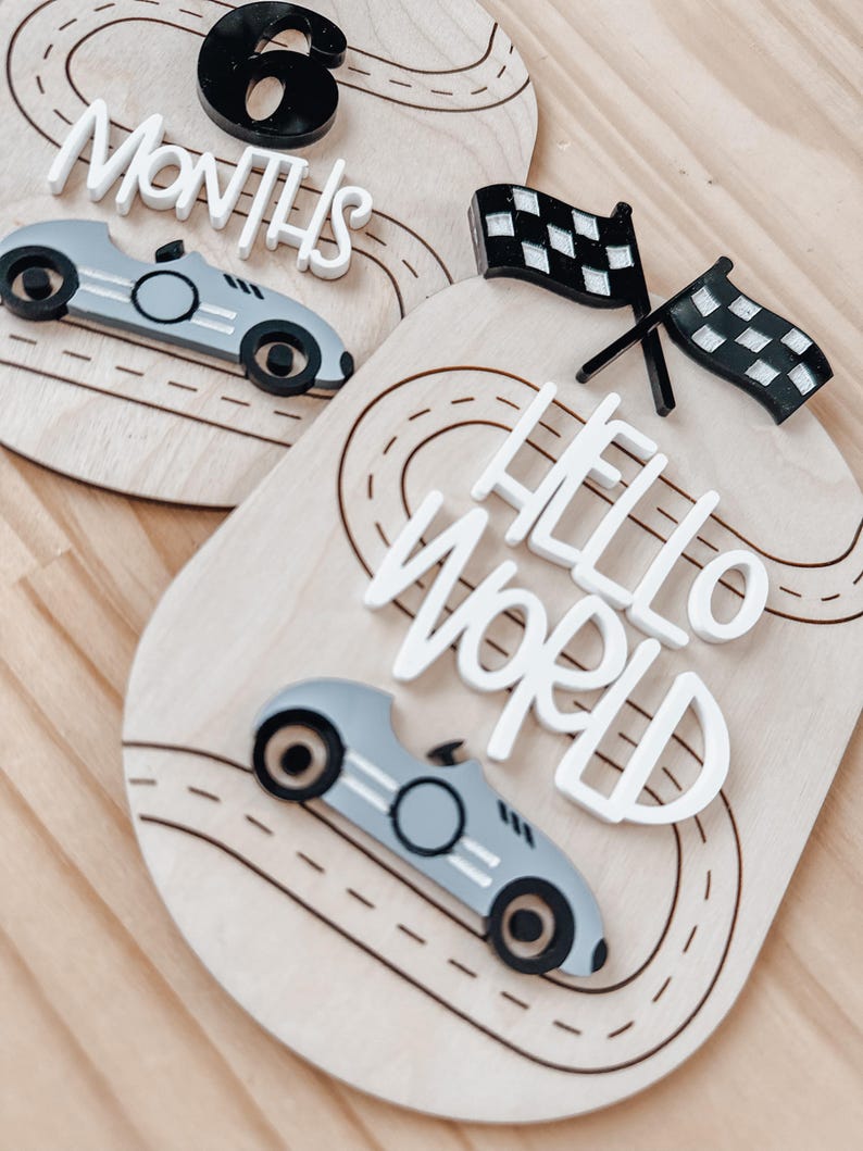 May include: Two wooden baby milestone signs with a race car theme. The first sign says "6 Months" and features a black number 6 and a grey race car. The second sign says "Hello World" and features a chequered flag and a grey race car.