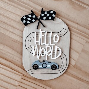 May include: Wooden sign featuring a race car, chequered flags, and the words "HELLO WORLD" on a black and white racetrack.