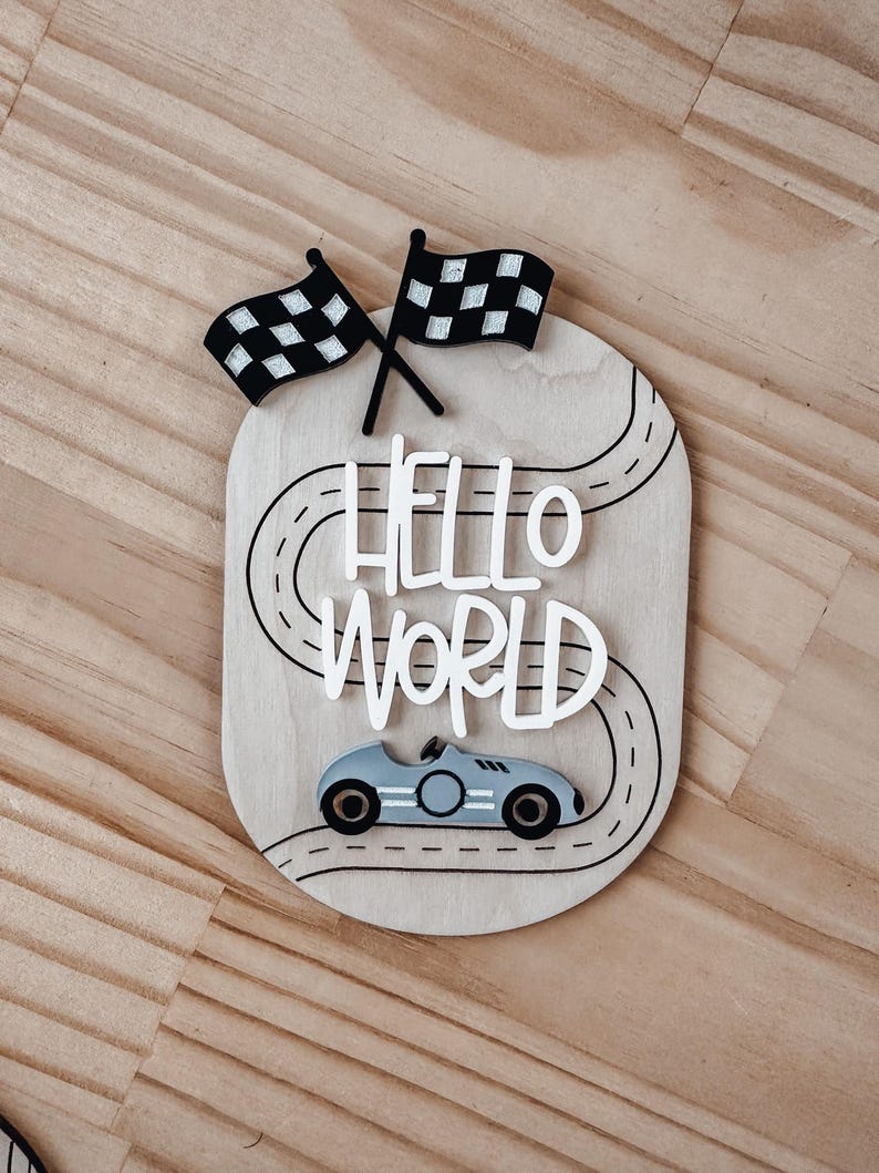 May include: Wooden sign featuring a race car, chequered flags, and the words "HELLO WORLD" on a black and white racetrack.