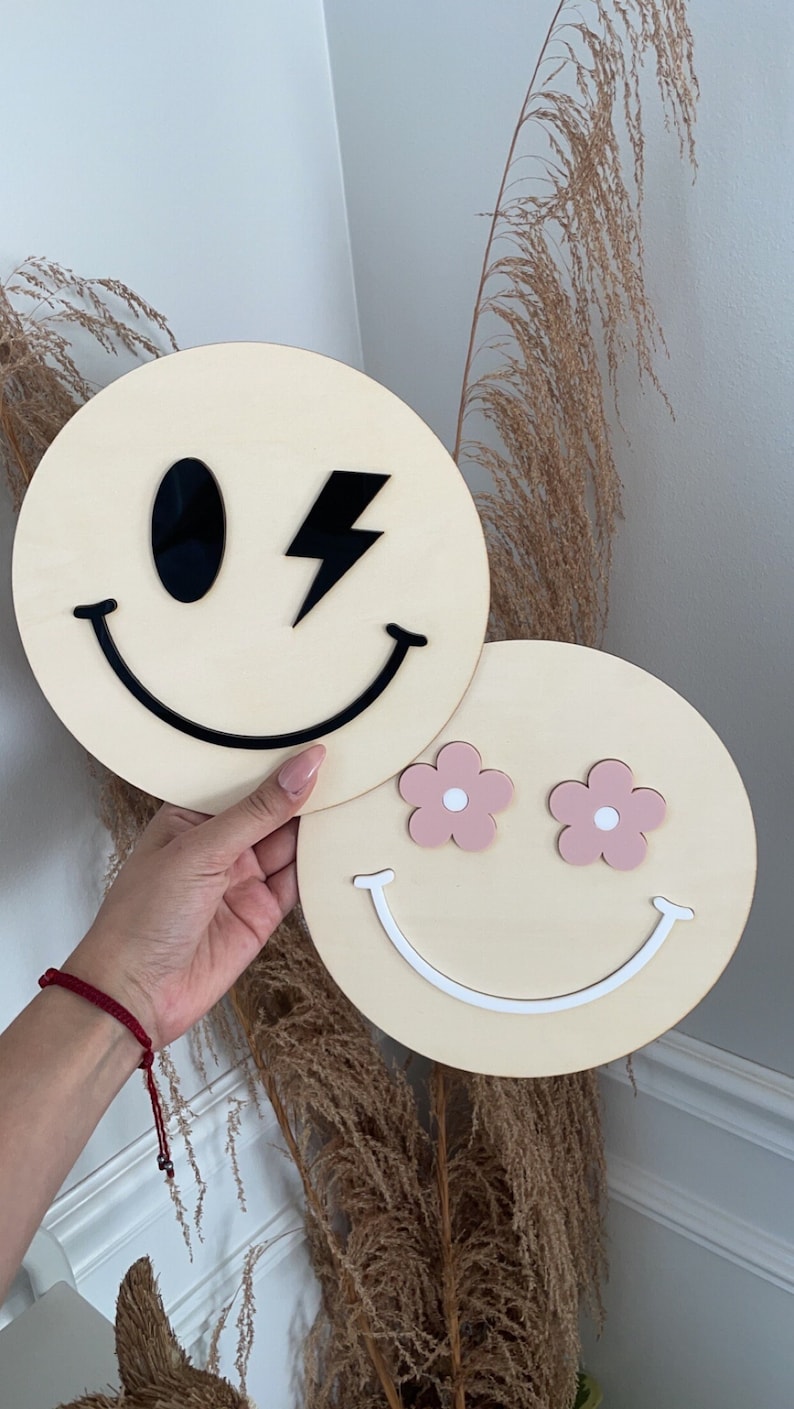 May include: Two wooden round smiley face wall decorations. One smiley face has a black lightning bolt and a black eye. The other smiley face has pink flower eyes with white centres.