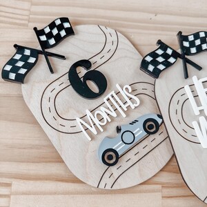 May include: Wooden milestone marker with a black number 6, the words "Months" and a black and white chequered flag. A grey race car is on a black and white race track.