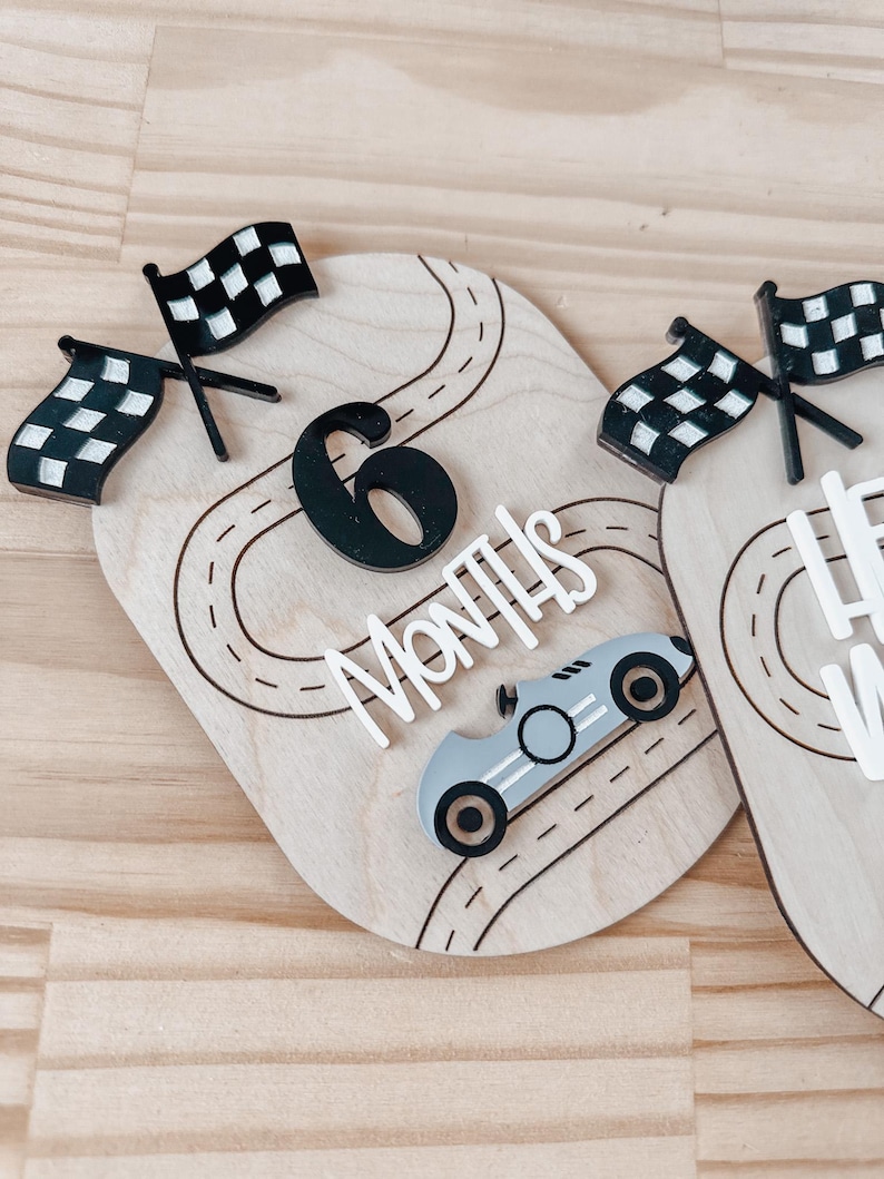 May include: Wooden milestone marker with a black number 6, the words "Months" and a black and white chequered flag. A grey race car is on a black and white race track.