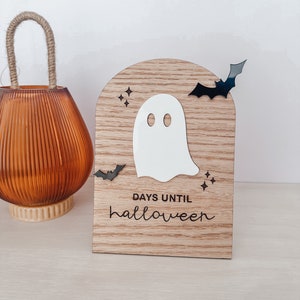 May include: Wooden Halloween countdown sign with a white ghost cutout, black bat accents, and the words "DAYS UNTIL halloween". The sign is light brown with a curved top. An orange glass jar with a rope handle is to the left.