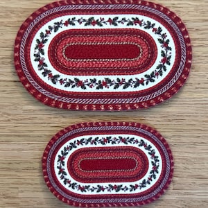 May include: Two oval braided rugs featuring a central solid red section surrounded by concentric rings of red, white, and a green holly leaf and red berry pattern. The rugs have a textured, woven appearance with a consistent color palette.