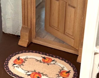 May include: A miniature oval rug with a beige centre and a dark purple border. The rug features a design of orange pumpkins and black crows with green vines. The rug is placed in front of a wooden door.