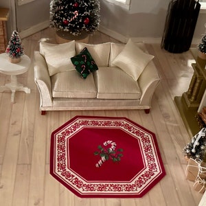 May include: A red and white octagonal rug with a candy cane design in the center. The rug is on a light wood floor in a room with a white couch and a small Christmas tree.