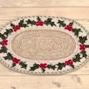 Op de afbeelding: Ovaal jute vloerkleed met een gevlochten rand. Het vloerkleed heeft een decoratieve rand van groene hulstbladeren en rode bessen, wat een feestelijk vakantiethema creëert. Het centrale gedeelte van het vloerkleed is naturel beige.