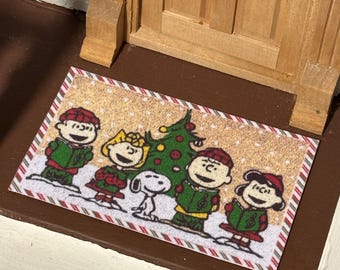 May include: A rectangular doormat featuring a festive scene with cartoon characters, a Christmas tree, and falling snow. The border has a red and white candy cane stripe design. The characters are wearing green and red outfits.