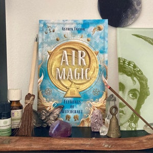 May include: A shelf display featuring a book titled "Air Magic: Elements of Witchcraft" with a blue and gold cover. Other items include crystals, incense, a small broom, essential oil bottles, and a decorative moon.