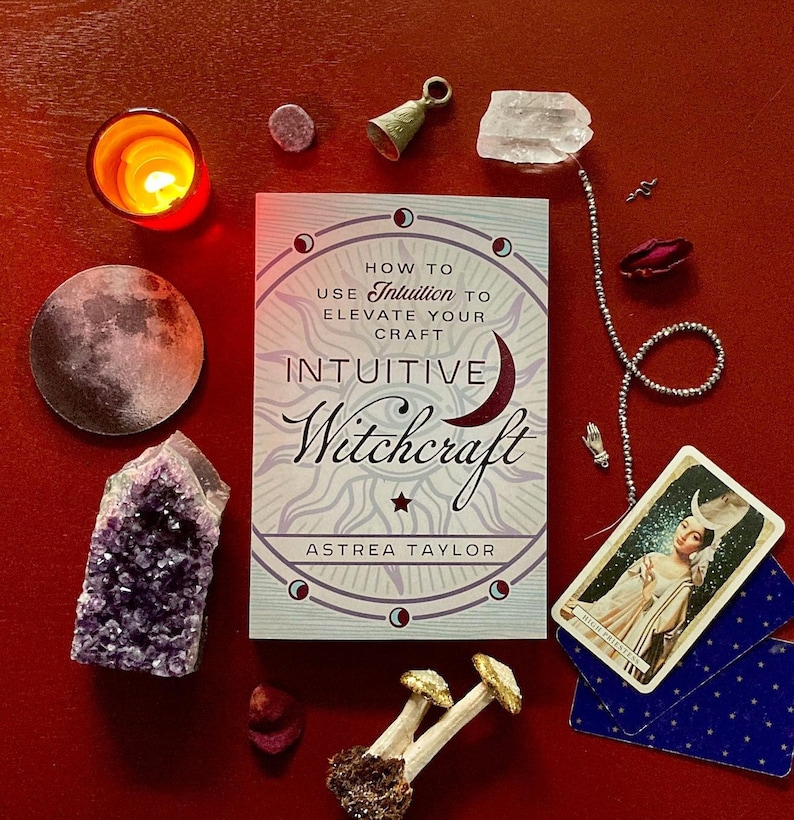 May include: A book titled "How to Use Intuition to Elevate Your Craft Intuitive Witchcraft" by Astrea Taylor. The book cover features a crescent moon, stars, and a starburst. The book is surrounded by various objects, including a candle, crystals, a bell, and a tarot card.