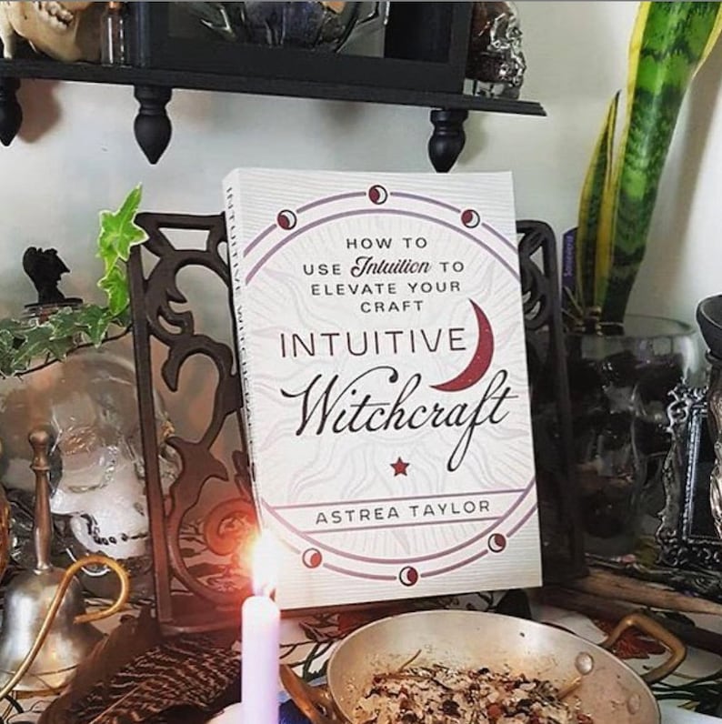 May include: A book titled "How to Use Intuition to Elevate Your Craft Intuitive Witchcraft" by Astrea Taylor. The book is on a wooden stand with a purple candle in front of it. There is a bowl with brown and red herbs in front of the candle.