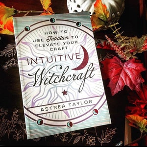 May include: A book titled "How to Use Intuition to Elevate Your Craft: Intuitive Witchcraft" by Astrea Taylor. The cover features a sun with a crescent moon and a star in the center. The book is on a dark surface with fall leaves.