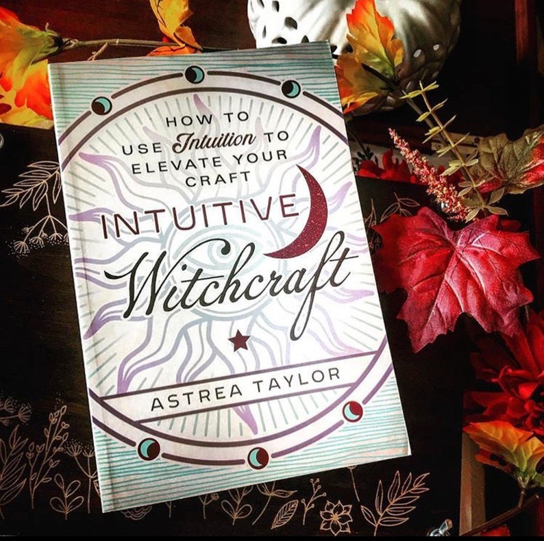 May include: A book titled "How to Use Intuition to Elevate Your Craft: Intuitive Witchcraft" by Astrea Taylor. The cover features a sun with a crescent moon and a star in the center. The book is on a dark surface with fall leaves.
