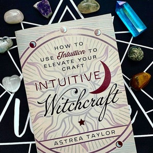 May include: A book titled "Intuitive Witchcraft: How to Use Intuition to Elevate Your Craft" by Astrea Taylor. The cover features a sun and moon design with a red crescent moon and a star.
