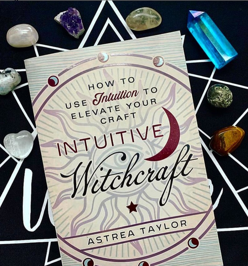 May include: A book titled "Intuitive Witchcraft: How to Use Intuition to Elevate Your Craft" by Astrea Taylor. The cover features a sun and moon design with a red crescent moon and a star.