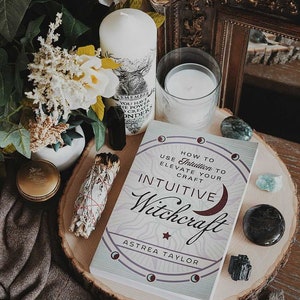 May include: A book titled "How to Use Intuition to Elevate Your Craft: Intuitive Witchcraft" by Astrea Taylor. The book cover features a crescent moon, stars, and a circle with a moon phase design. The book is on a wooden slice with crystals and a white candle.