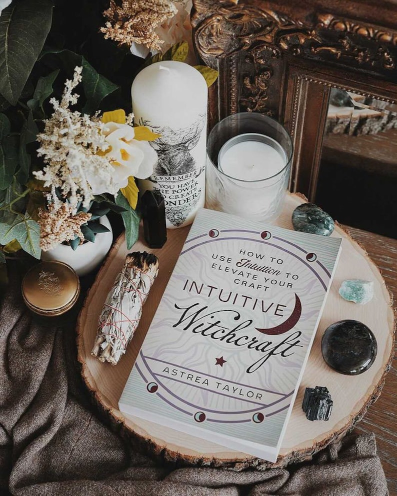 May include: A book titled "How to Use Intuition to Elevate Your Craft: Intuitive Witchcraft" by Astrea Taylor. The book cover features a crescent moon, stars, and a circle with a moon phase design. The book is on a wooden slice with crystals and a white candle.