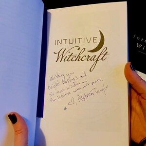 May include: A white book with the title "Intuitive Witchcraft" written in black cursive font. A crescent moon symbol is next to the title. The book is signed by the author, "Astren Taylor", with a message wishing the reader bright blessings and wisdom on their intuitive witchcraft path.