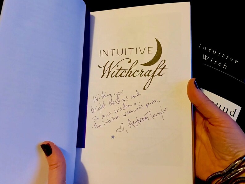 May include: A white book with the title "Intuitive Witchcraft" written in black cursive font. A crescent moon symbol is next to the title. The book is signed by the author, "Astren Taylor", with a message wishing the reader bright blessings and wisdom on their intuitive witchcraft path.