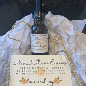 May include: A small, clear glass bottle with a black dropper top filled with a dark liquid. The bottle is labeled "Arnica Flower Essence" and has the text "I let go my past & I accept to trust & re-connect with my physical & spiritual self love and joy" on a white card with yellow flower illustrations.