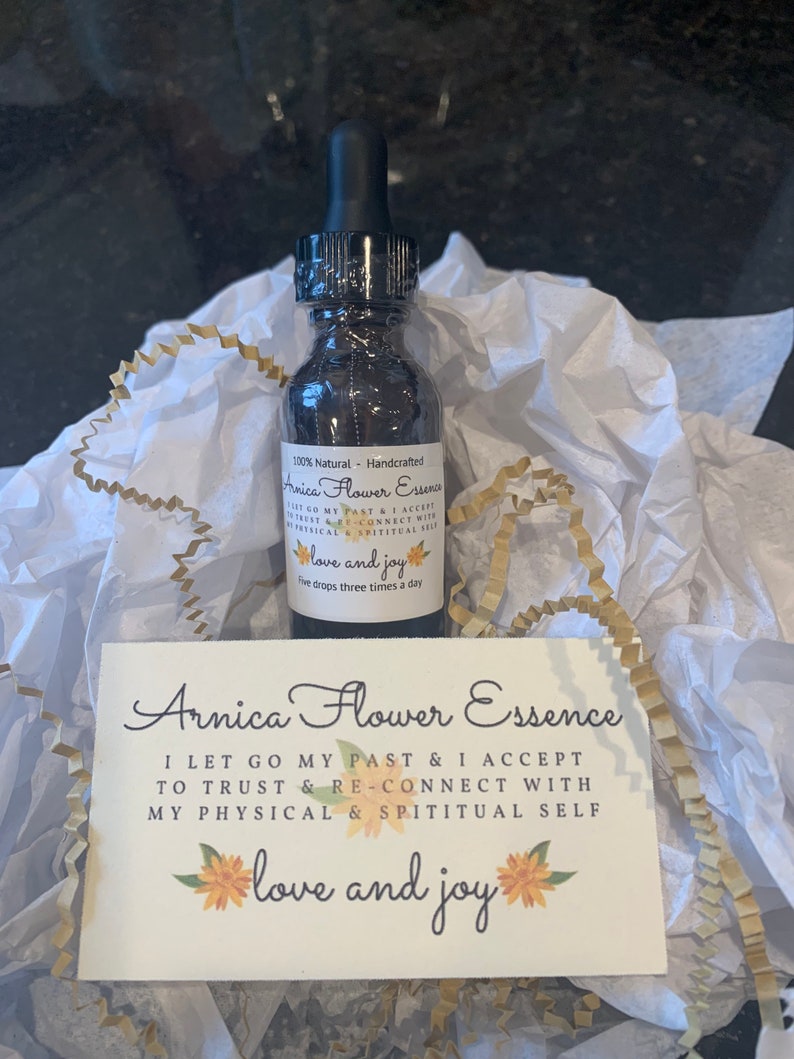 May include: A small, clear glass bottle with a black dropper top filled with a dark liquid. The bottle is labeled "Arnica Flower Essence" and has the text "I let go my past & I accept to trust & re-connect with my physical & spiritual self love and joy" on a white card with yellow flower illustrations.