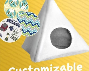 May include: A white, triangular pet bed with a circular opening. Three circular fabric swatches show various patterns, including bunnies, zigzags, and camping-themed designs. The word "Customizable" is printed in white at the bottom.
