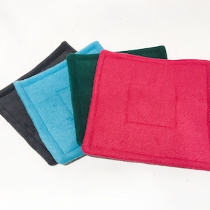 May include: Four square fabric coasters in different colours: black, blue, green, and red. The coasters are made of a soft, fuzzy material and have a stitched border.