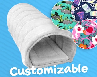 May include: A white, plush, customizable pet bed with a rectangular interior. The image includes three circular fabric swatches with various patterns: hippos in a jungle, space-themed designs, and floral patterns. The word "Customizable" is written in white.