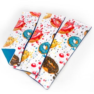 May include: Three long, rectangular fabric heat packs with a donut and sprinkles pattern. The heat packs are blue on the back and white with colorful donuts on the front.