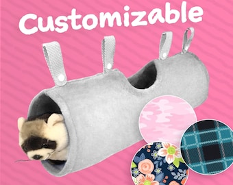 May include: A gray, customizable pet tunnel with a plush raccoon inside. The tunnel has three hanging loops and is shown with three fabric pattern options: pink camouflage, floral, and plaid. The word "Customizable" is in white text.