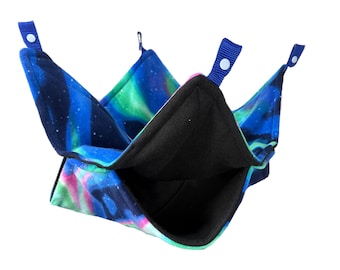May include: A small pet hammock with a vibrant aurora borealis pattern in blue, green, and pink. The interior is black, and the hammock has blue fabric loops with white snaps for hanging. This cozy accessory is designed for small pets.