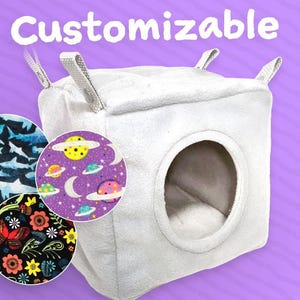 May include: A white, cube-shaped pet bed with a circular opening. The word "Customizable" is above the bed. Three circular fabric swatches show various patterns: sharks, planets, and flowers. The bed has four grey fabric loops.