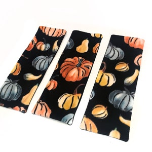 Può includere: Tre segnalibri in tessuto nero con un motivo a zucca e zucca. I segnalibri sono rettangolari e hanno un bordo nero.