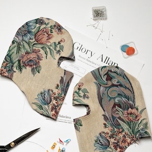 May include: Two balaclava mask cutouts with a floral tapestry design in beige, green, and burgundy. The image includes sewing supplies like pins, clips, and scissors, along with a pattern sheet that reads "Glory Allan Woven Balaclava".