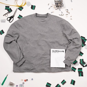 May include: A grey long-sleeved shirt with a white booklet that says "GLORYALLAN" on the cover. The shirt is surrounded by sewing supplies, including scissors, pins, and fabric scraps.