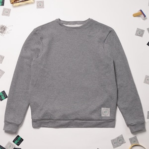 May include: A gray sweatshirt with a small white label on the bottom right side. The sweatshirt is laid flat on a white surface with various small green and black stickers scattered around it.
