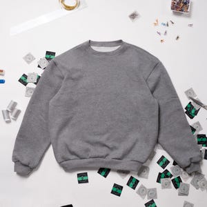May include: A gray sweatshirt with long sleeves and a crew neck. The sweatshirt is lying flat on a white surface with various craft supplies scattered around it.