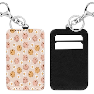 May include: A cardholder keychain with a smiley face and daisy pattern on a light pink background. The other side is solid black with two card slots. The keychain has a silver metal clasp.