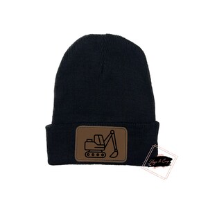 May include: A black knit beanie with a brown rectangular patch featuring a line drawing of a construction excavator. The beanie has a folded brim and is set against a white background. The patch is centered on the front of the beanie.