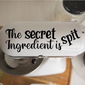 May include: A white stand mixer with the words "The secret ingredient is spit" in black script. The mixer is on a wooden surface, with other kitchen items visible in the background.