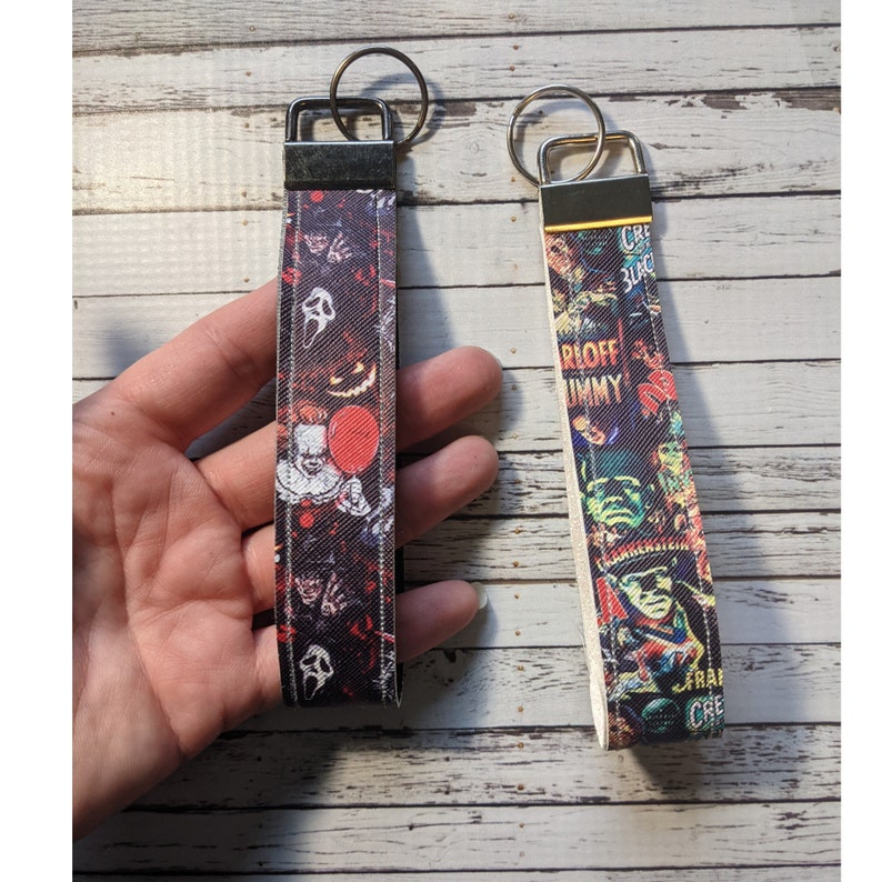 May include: Two colorful keychains with horror movie themed designs. The keychain on the left features a black and red design with characters from the movie "IT" and "Scream". The keychain on the right features a white and colorful design with characters from classic horror movies like "Creature from the Black Lagoon" and "The Mummy".