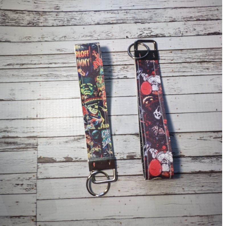 May include: Two colorful keychains with metal rings. One keychain features a black and white image of a monster with the text "Frankenstein, Creature from the Black Lagoon". The other keychain features a red and white image of a clown with the text "Pennywise".