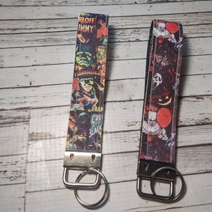 May include: Two colorful keychains with silver key rings. The keychain on the left features a collage of horror movie posters with the text "Frankenstein" and "Creature from the Black Lagoon". The keychain on the right features a collage of horror movie characters including Pennywise the clown from "IT".