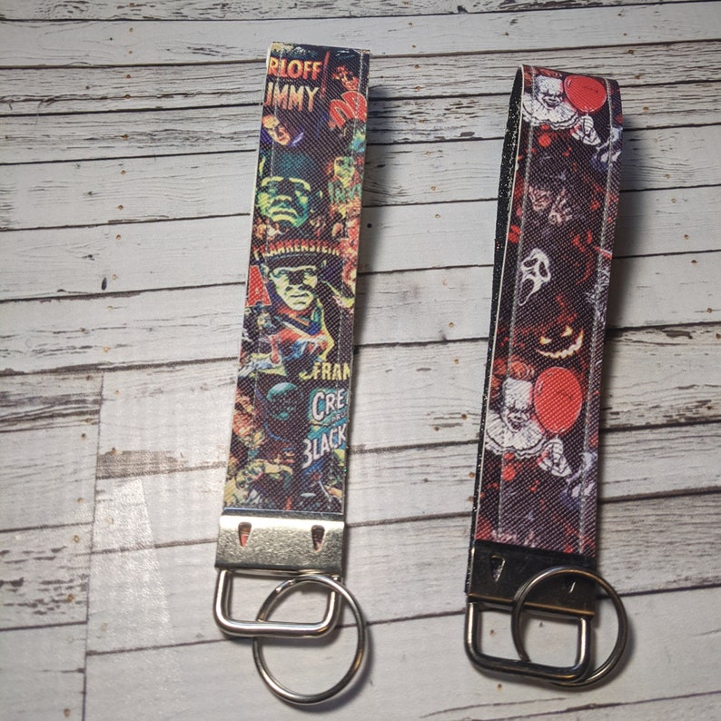 May include: Two colorful keychains with silver key rings. The keychain on the left features a collage of horror movie posters with the text "Frankenstein" and "Creature from the Black Lagoon". The keychain on the right features a collage of horror movie characters including Pennywise the clown from "IT".