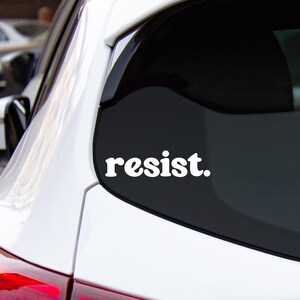 Könnte beinhalten: Weißer Vinyl-Aufkleber mit dem Wort "resist." auf schwarzem Hintergrund. Der Aufkleber befindet sich auf der Heckscheibe eines weißen Autos.