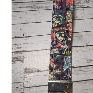 May include: A white fabric keychain with a colorful print of classic horror movie posters. The print features images of Frankenstein, Dracula, and other monsters. The keychain has a silver metal clasp.
